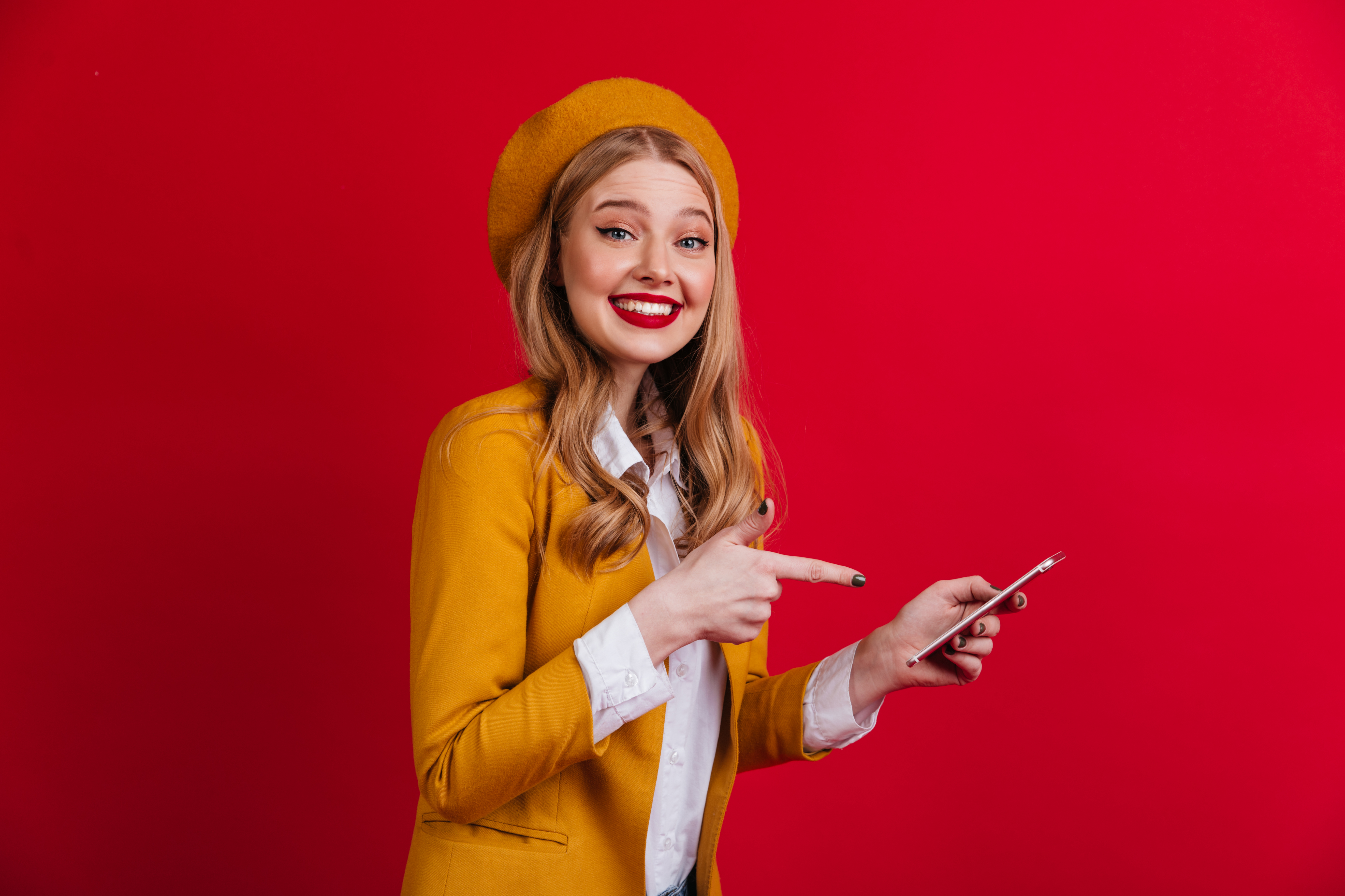 cheerful-french-lady-pointing-with-finger-smartphone-smiling-blonde-girl-beret-standing-red-wall
